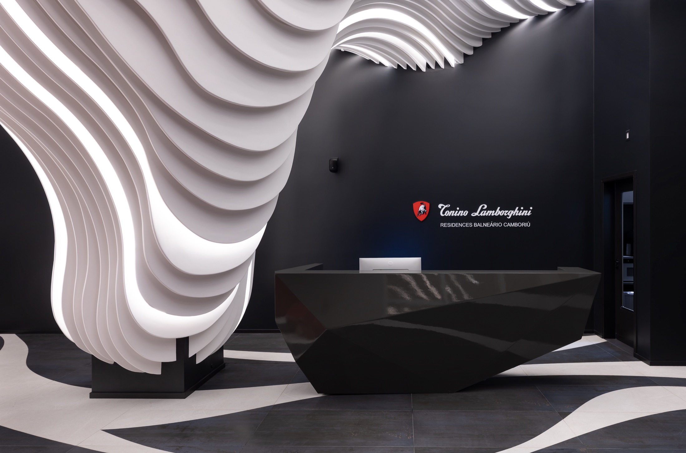 Fotografía real del Hall de Entrada entregado, mostrando la escultura paramétrica blanca suspendida, el mostrador facetado en piedra negra y el escudo Tonino Lamborghini