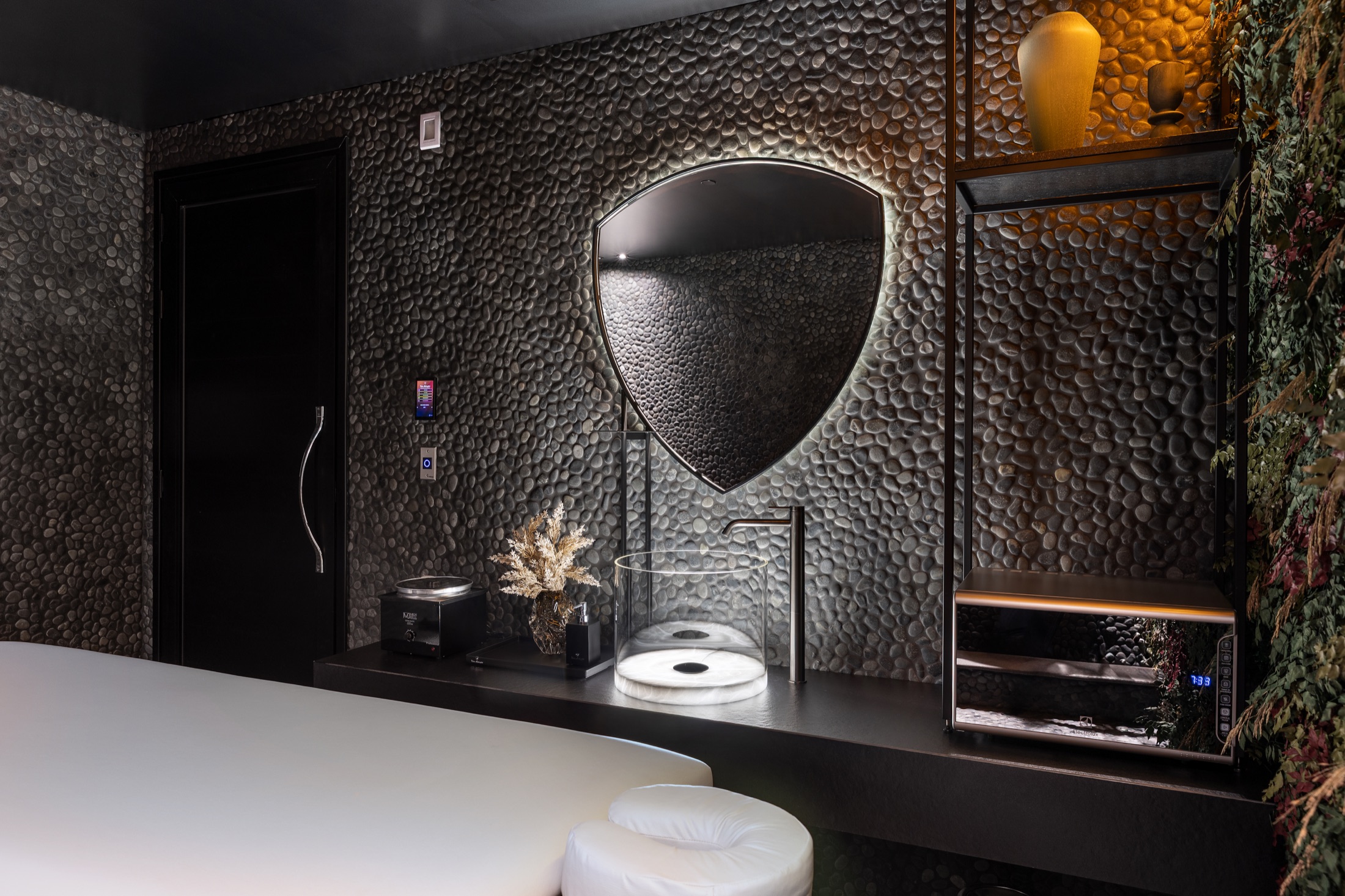 Fotografía real de la sala de masajes con pared en guijarros negros naturales, jardín vertical y espejo retroiluminado con la forma del escudo Tonino Lamborghini